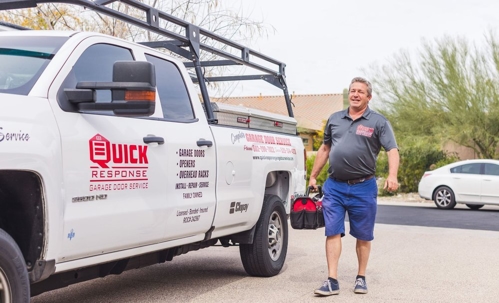 A garage door repairman is walking towards a customer's house with a toolbox in hand, next to his company truck.